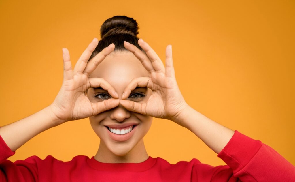 Portrait of a smiling woman making a playful eye gesture with her hands against an orange background.